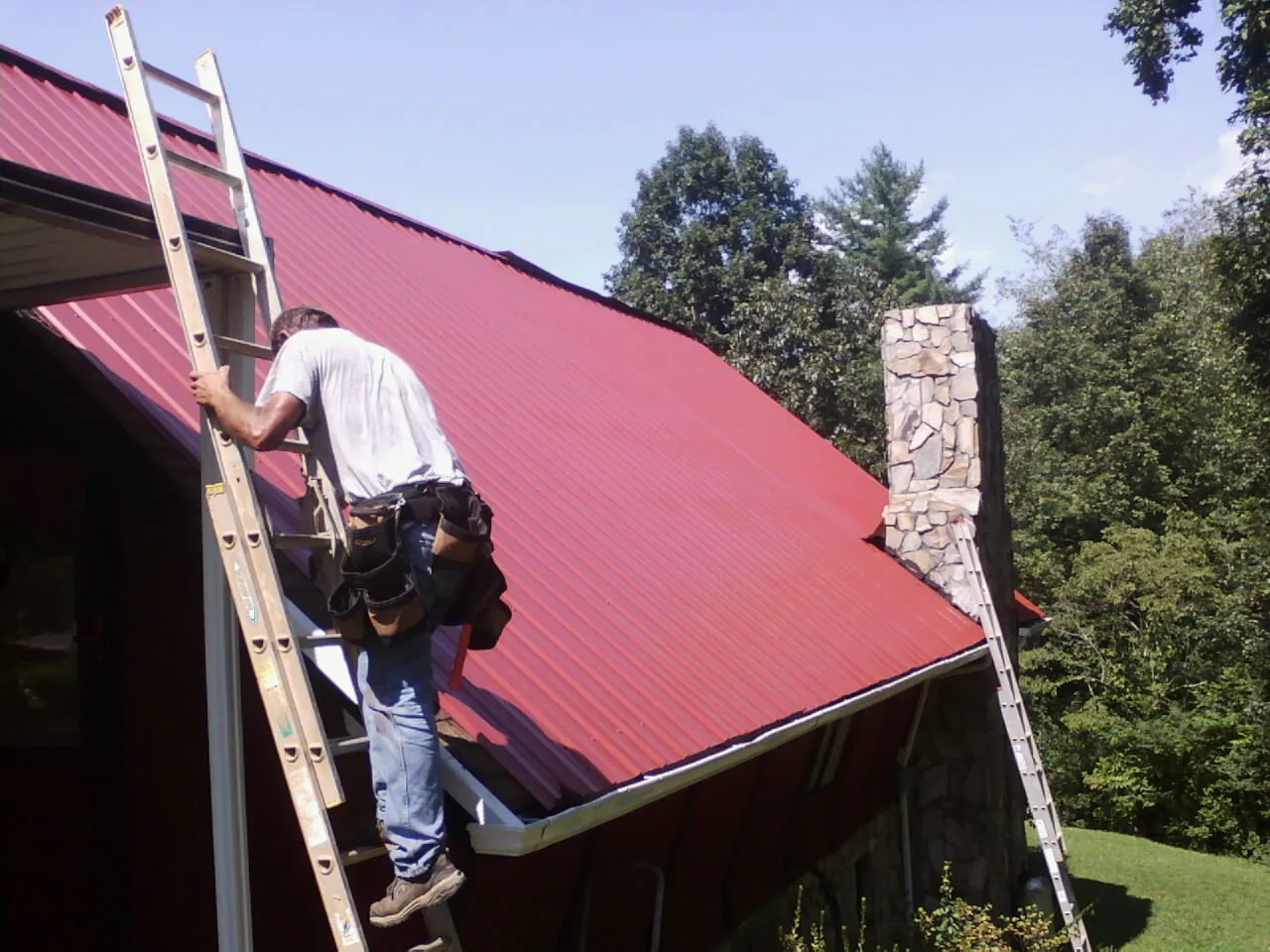 Roofer on a ladder beside a red metal roof and stone chimney — Soffit & Fascia Repair by Roofing Brothers in Corvallis